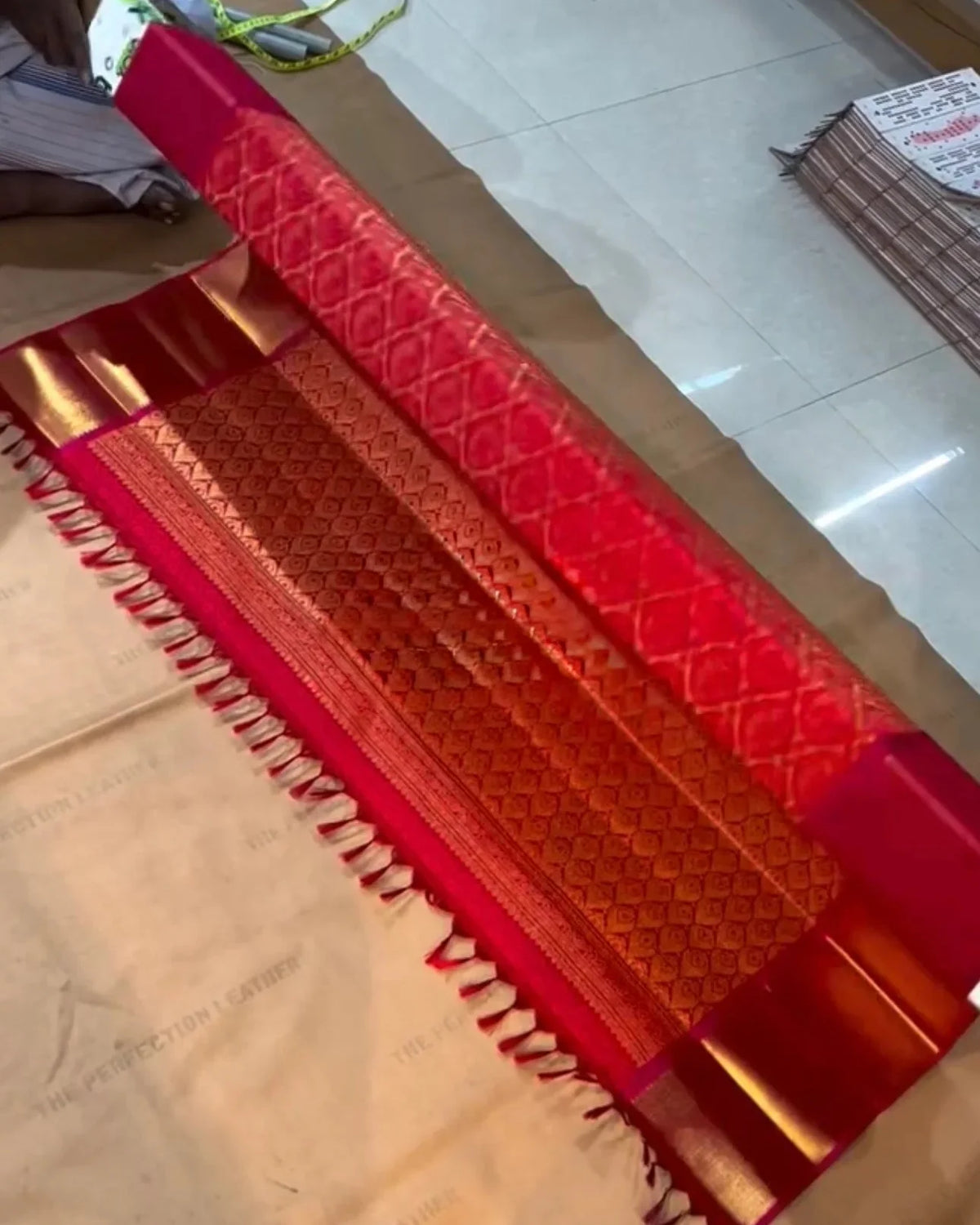 a red and gold zari woven saree with a patterned border and white fringe, laid out on a light-colored floor.