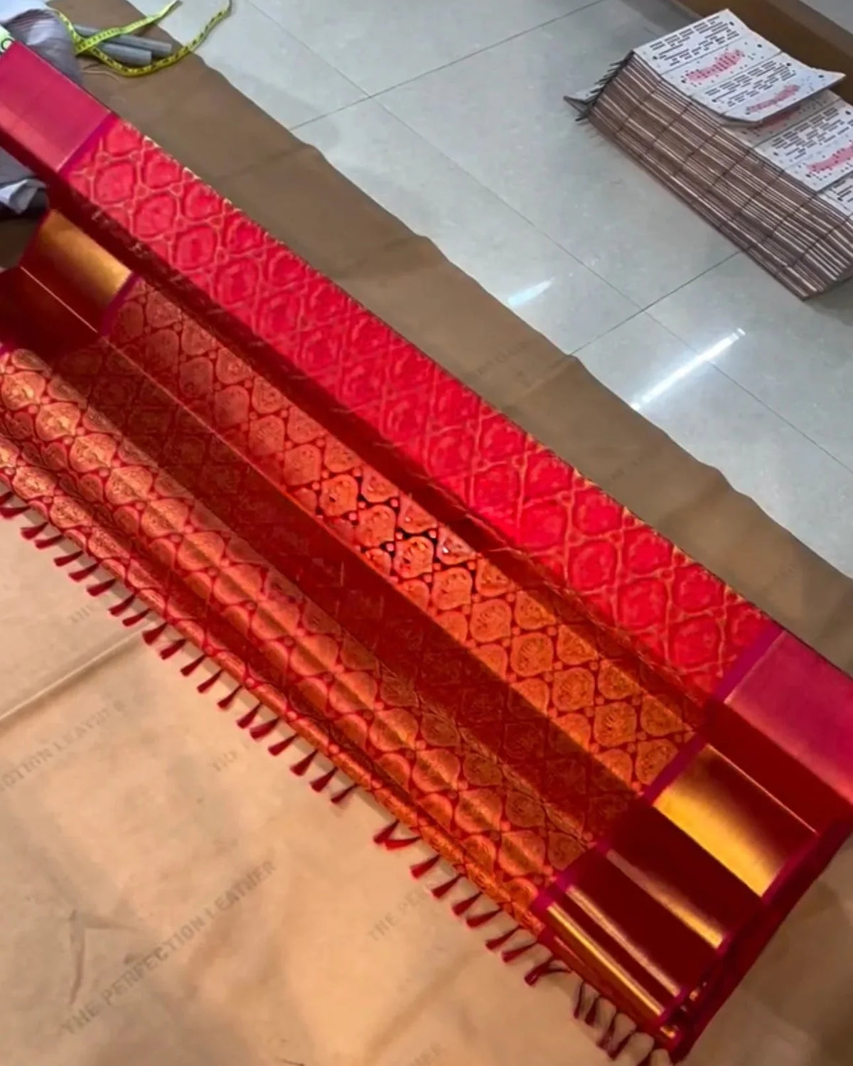 A red and gold patterned leather belt with fringe detailing, laid diagonally across a light-colored floor.
