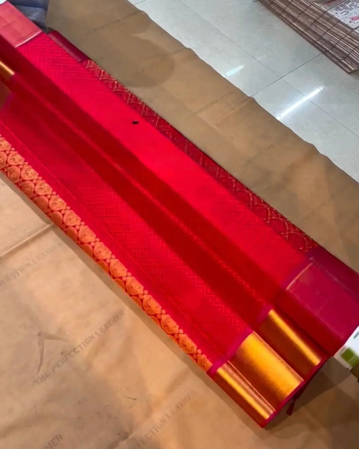 a close-up of a red fabric or leather material roll with gold and orange woven borders, laid diagonally on a cardboard surface in an indoor setting.