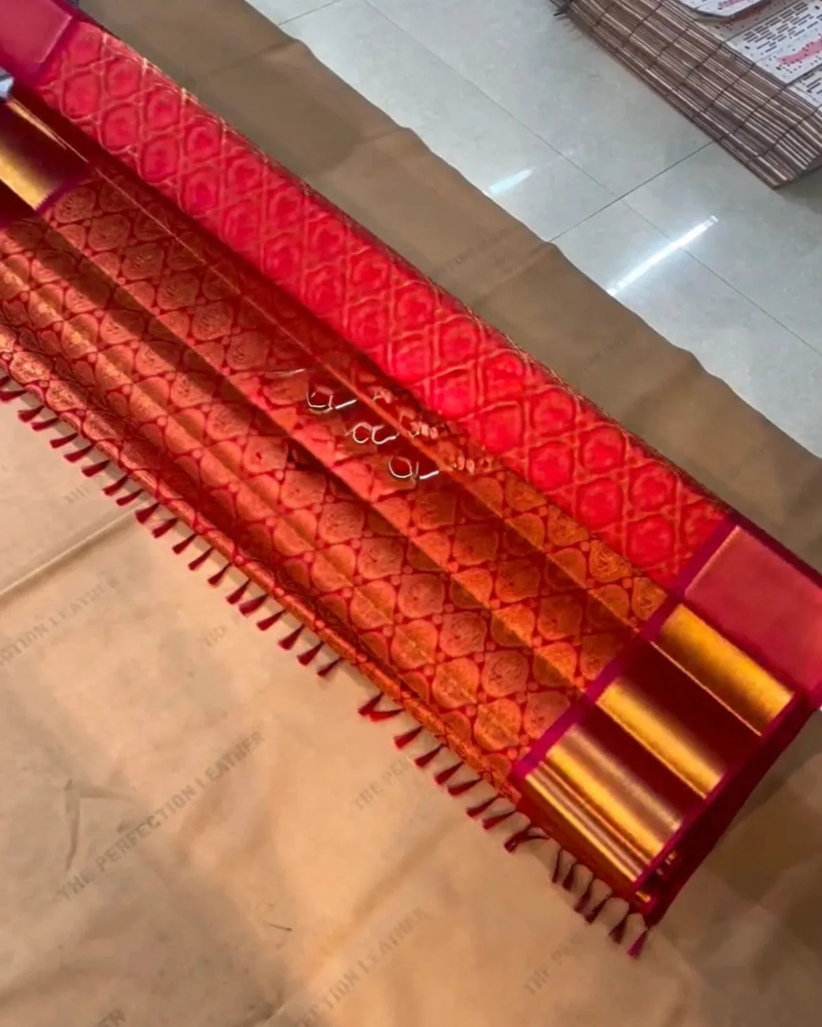 A close-up of a red and gold patterned fabric, likely a saree or scarf, laid out on a light-colored floor with a stack of similar fabrics in the background.